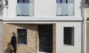 Fachada de casa con ladrillo visto y pared blanca. Una puerta oscura y ventanas con rejas. Ideas para como construir una casa barata en España.