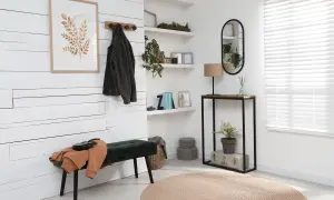 Un recibidor estrecho con un banco verde, perchero con chaqueta, cuadro de hojas y consola con espejo y lámpara.
