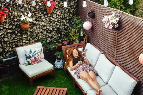 Mujer relajándose en un patio decorado para **decorar patios pequeños con poco dinero**. Flores, luces y muebles crean un oasis acogedor.