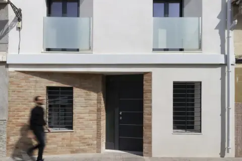Fachada de casa con ladrillo visto y pared blanca. Una puerta oscura y ventanas con rejas. Ideas para como construir una casa barata en España.