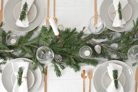 Sencillo decoracion mesa navidad con ramas de pino, piñas y velas. Vajilla gris, servilletas blancas con detalle de romero y cubertería dorada.