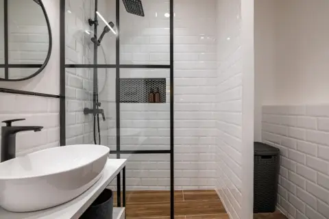 Moderno murete baño con ducha de cristal, lavabo blanco y grifería negra. Azulejos blancos y suelo de madera.