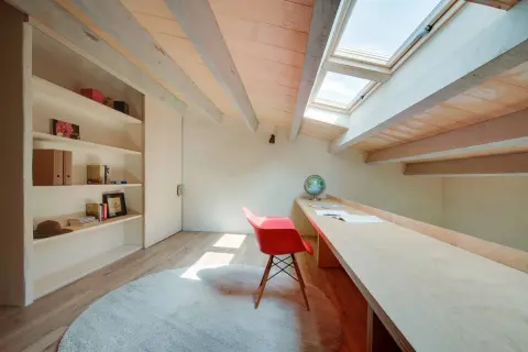 Un pequeño patio de luz interior baña con sol un escritorio de madera, una silla roja y una estantería.