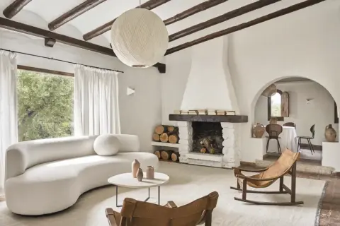 Salón acogedor con sofá curvo blanco, chimenea de piedra y mecedora de cuero. Un ejemplo de casas de campo sencillas y bonitas.