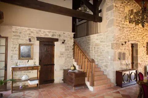 Interior de una casa antigua con paredes de piedra, escaleras de madera, un baúl y muebles rústicos.