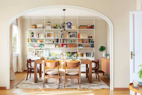Comedor con decoración estilo nórdico, mesa de madera, sillas de diseño y estanterías blancas repletas de libros y objetos decorativos.