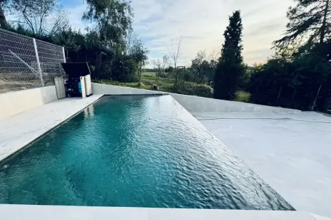 Una moderna piscina infinita con agua turquesa, rodeada de un área de concreto blanco y vegetación exuberante. El cielo azul con nubes blancas completa la escena.