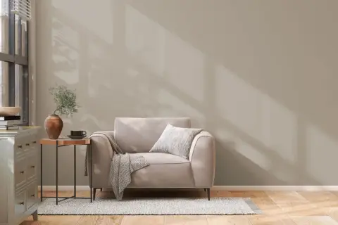 Un sillón beige con manta y cojín, junto a una mesita auxiliar con jarrón y taza. La pared tiene unos colores de pintura neutros, con sombras de luz.