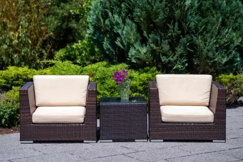 Dos sillones y una mesita de centro de ratán, perfectos para el mantenimiento de muebles de exterior. Flores moradas en un jarrón.