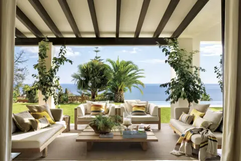 Cómodos porches de entrada principal con sofás de madera, cojines y mantas, con vistas al mar y palmeras.
