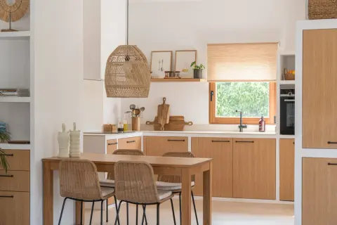Cocina estilo mediterráneo con mesa de madera, sillas de mimbre y armarios de roble. Lámpara de mimbre y ventana con vistas a la vegetación.