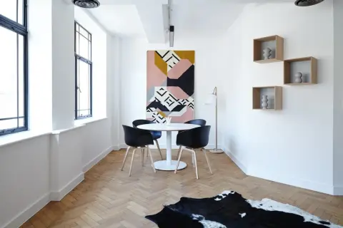 Un comedor moderno con mesa redonda, sillas negras y una alfombra geométrica. Las estanterías flotantes y las ventanas grandes dan la sensación de casas pequeñas y acogedoras.
