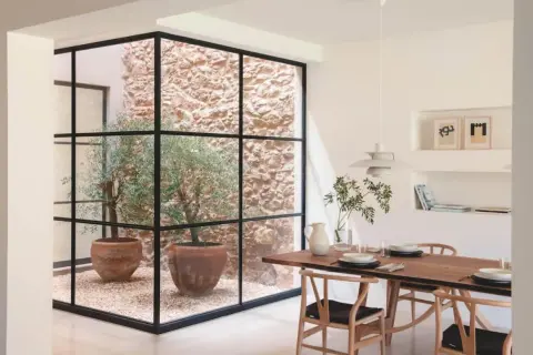 Un comedor moderno con una mesa de madera y sillas de diseño, junto a un patio de luces con olivos en macetas y paredes de piedra.