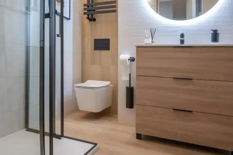 Modernos baños blancos y madera clara, con ducha de cristal, inodoro suspendido y mueble de cajones.