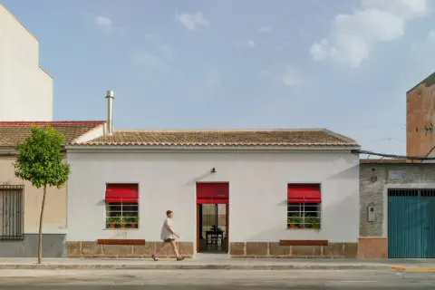 Fachada casa pueblo moderna con persianas rojas vibrantes y un hombre corriendo.