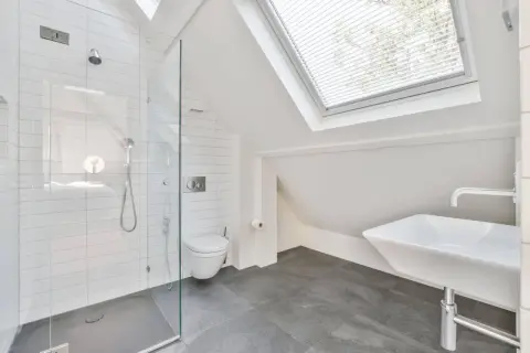 Moderno baño en buhardilla con ducha de cristal, inodoro y lavabo bajo una claraboya.