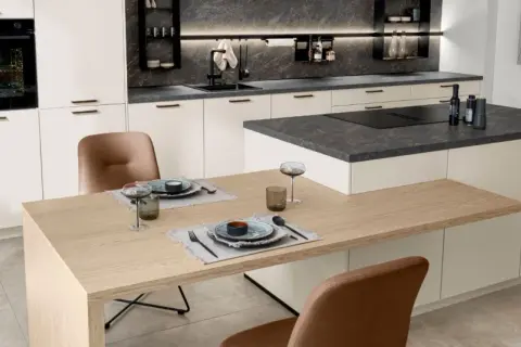 Cocina moderna con encimeras de piedra oscura y gabinetes blancos. La mesa de madera clara y las sillas de cuero marrón complementan los materiales para cocinas modernas.