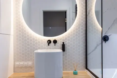 Moderno baño con lavabo blanco, espejo redondo iluminado y azulejos hexagonales. Ideal para tu presupuesto baño.