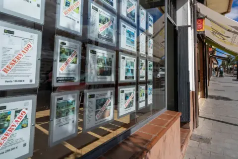 Escaparate de inmobiliaria con anuncios de venta de casas, muchos marcados como "RESERVADO". Se vislumbra una casa señorial en uno de los carteles.