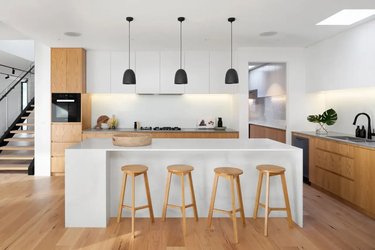 Moderna cocina blanca y madera con isla central, taburetes de madera y escaleras de diseño.