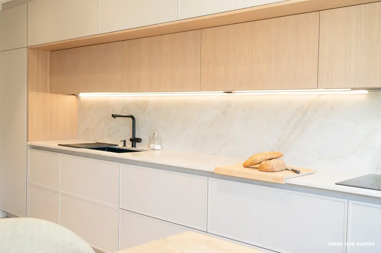 Cocina blanca y gris con armarios de madera clara, encimera de m&aacute;rmol y grifo negro.