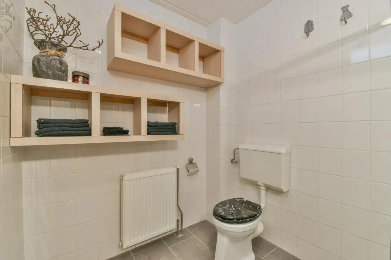 Estanterías de madera con toallas y un jarrón decorativo en estos baños pequeños. Un radiador blanco y un inodoro completan la escena.