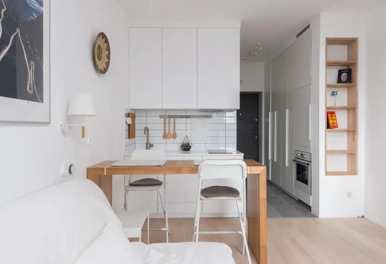 Cocina y sala de estar integradas en a casa pequena. Muebles blancos, barra de madera y estantería de roble.
