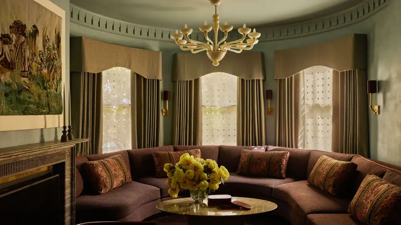 Sofá curvo marrón con cojines decorativos, mesa redonda con flores amarillas y un tapiz en la pared, todo con un toque de art deco.