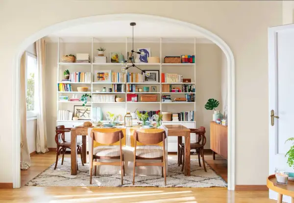 Comedor con decoración estilo nórdico, mesa de madera, sillas de diseño y estanterías blancas repletas de libros y objetos decorativos.
