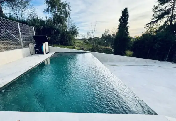 Una moderna piscina infinita con agua turquesa, rodeada de un área de concreto blanco y vegetación exuberante. El cielo azul con nubes blancas completa la escena.