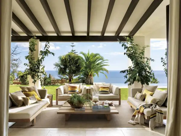 Cómodos porches de entrada principal con sofás de madera, cojines y mantas, con vistas al mar y palmeras.