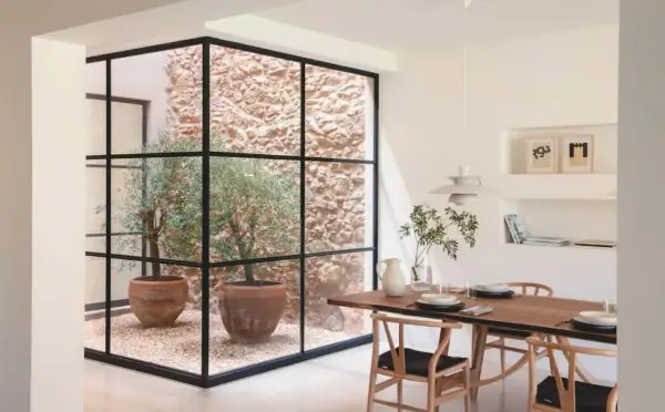 Un comedor moderno con una mesa de madera y sillas de diseño, junto a un patio de luces con olivos en macetas y paredes de piedra.